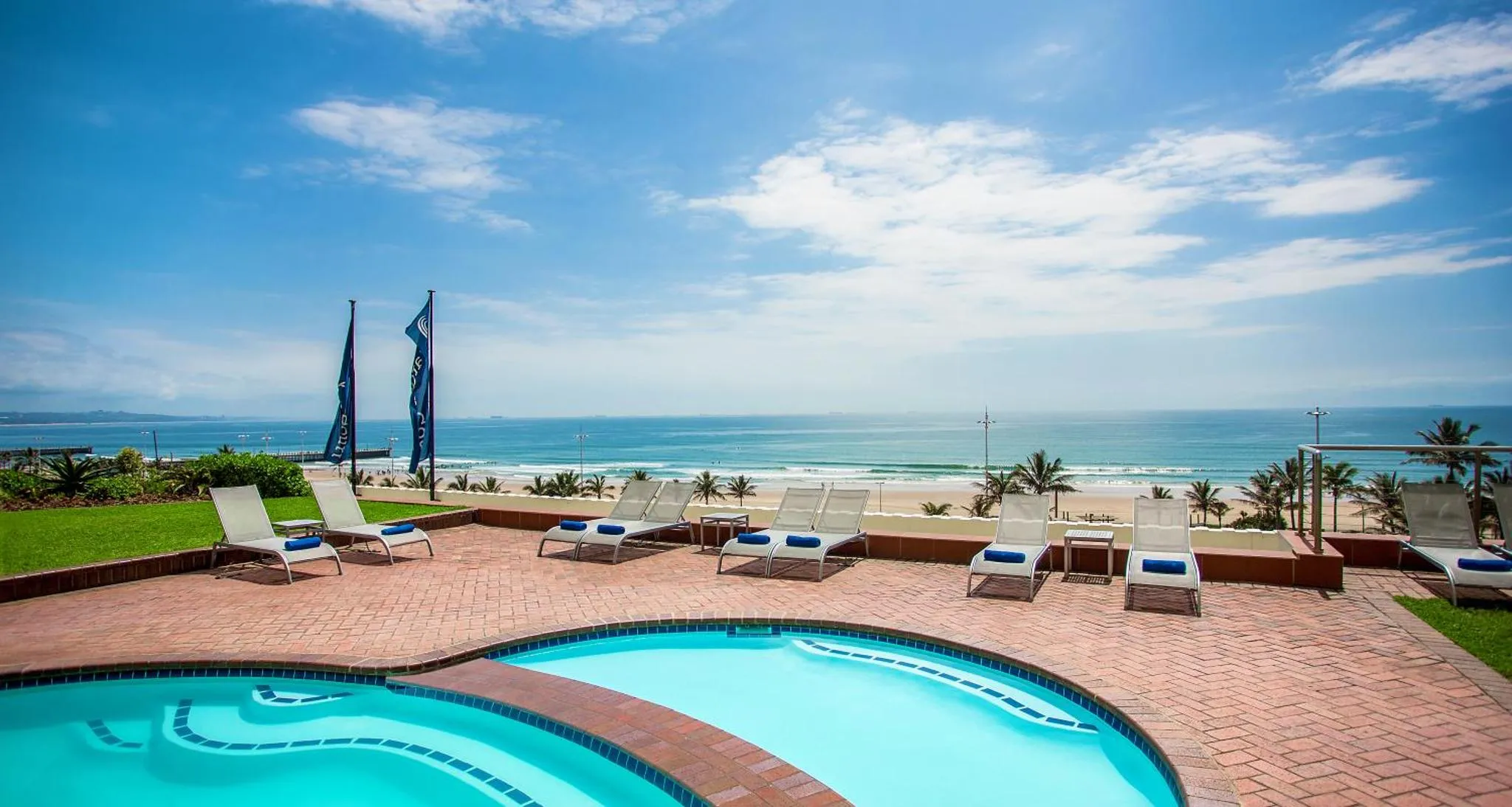 Transats bleus alignés sur une terrasse ensoleillée avec piscine et vue sur la mer.