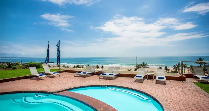Transats bleus alignés sur une terrasse ensoleillée avec piscine et vue sur la mer.