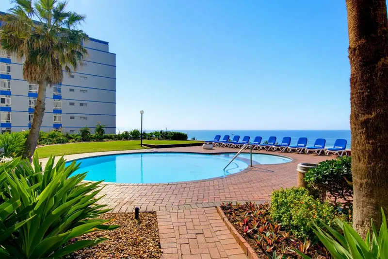 Une piscine rectangulaire bordée de chaises longues se trouve devant un bâtiment et la mer.