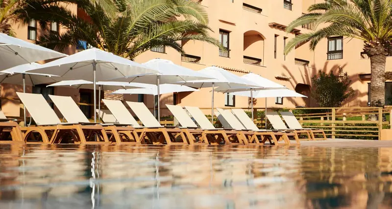 Transats blancs disposés au bord d'une piscine turquoise, sous des parasols.