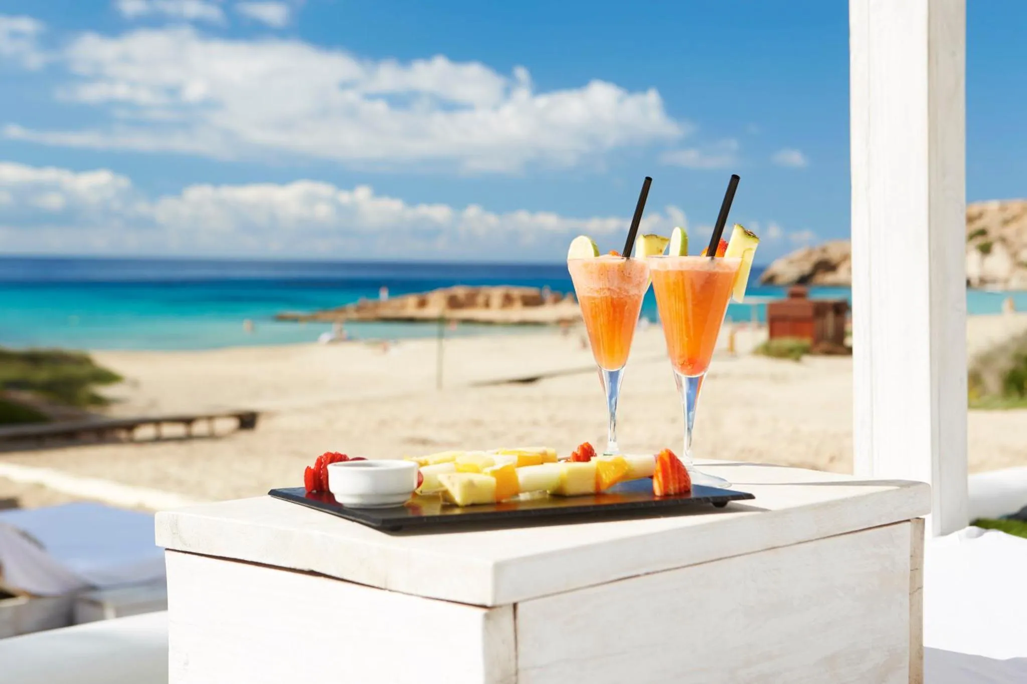 Deux cocktails colorés et des fruits frais sont présentés sur une table blanche face à une plage de sable clair et une mer turquoise.