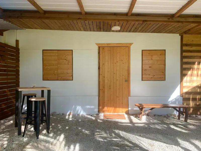 Une terrasse ombragée donne sur une façade blanche avec une porte en bois.