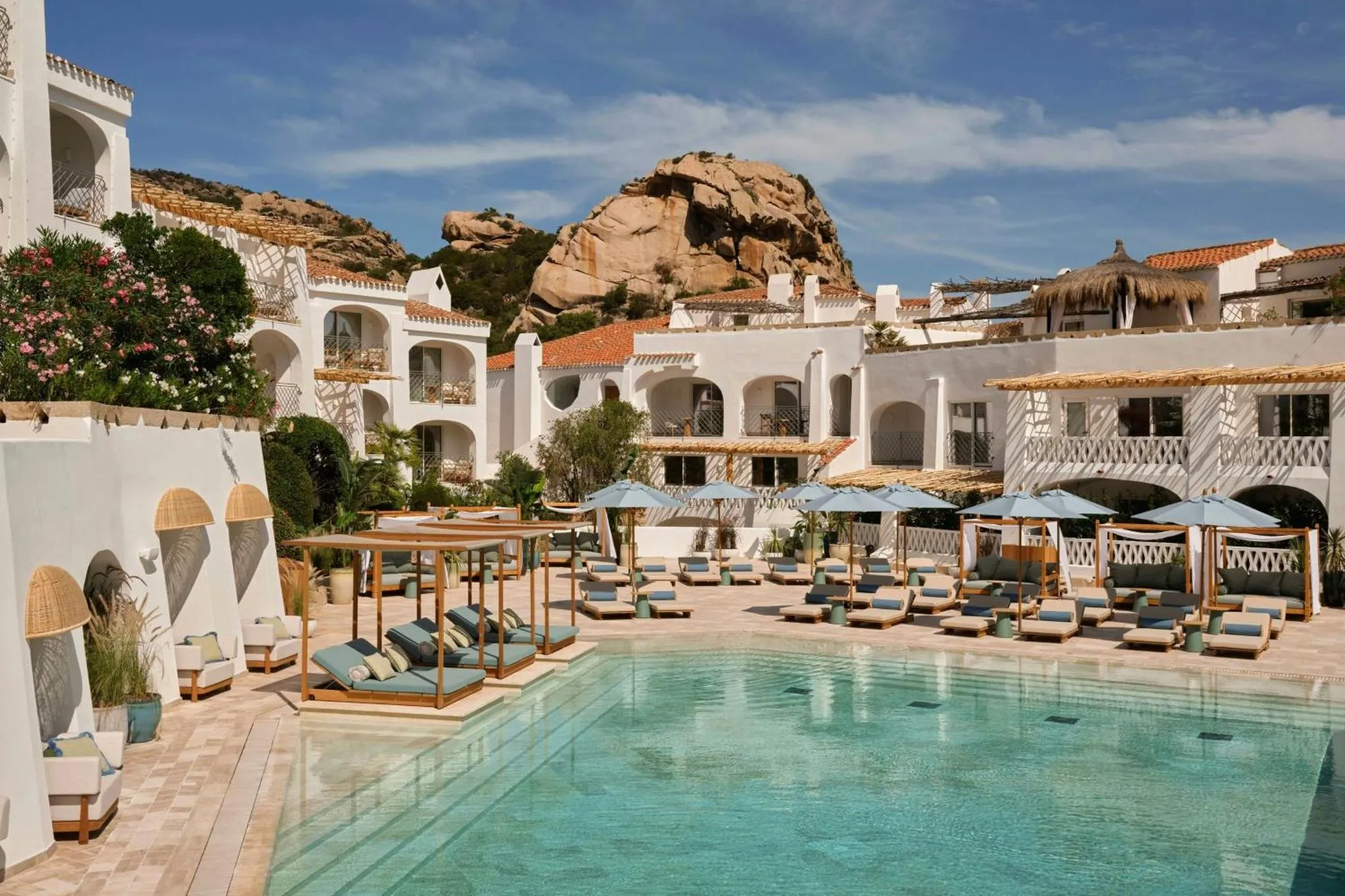 Une piscine turquoise bordée de transats et de parasols, devant un bâtiment blanc.