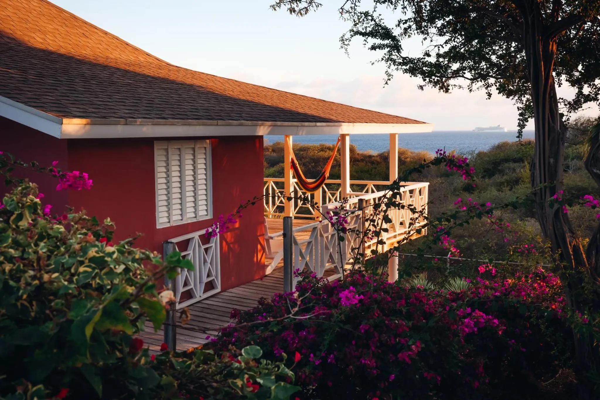 Une maison rouge avec une terrasse en bois et un hamac face à la mer.