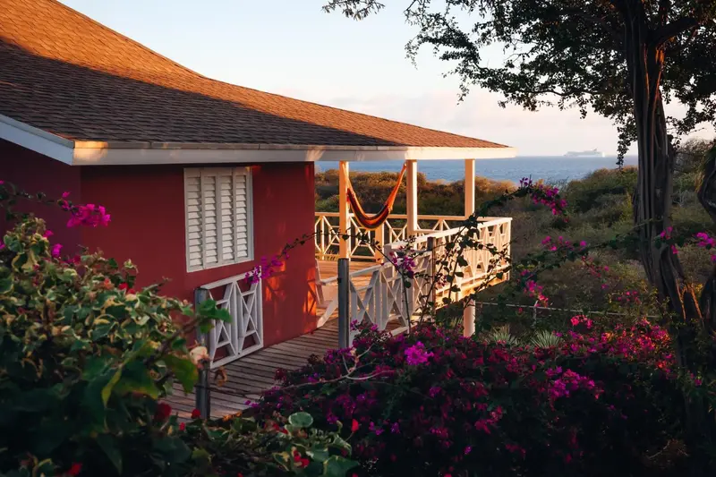 Une maison rouge avec une terrasse en bois et un hamac face à la mer.