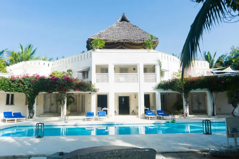 Bâtiment blanc à deux étages avec une piscine au premier plan et une végétation luxuriante.
