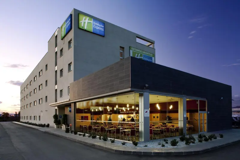 Façade d'un bâtiment moderne avec une entrée illuminée et des tables de restaurant visibles à travers les fenêtres.