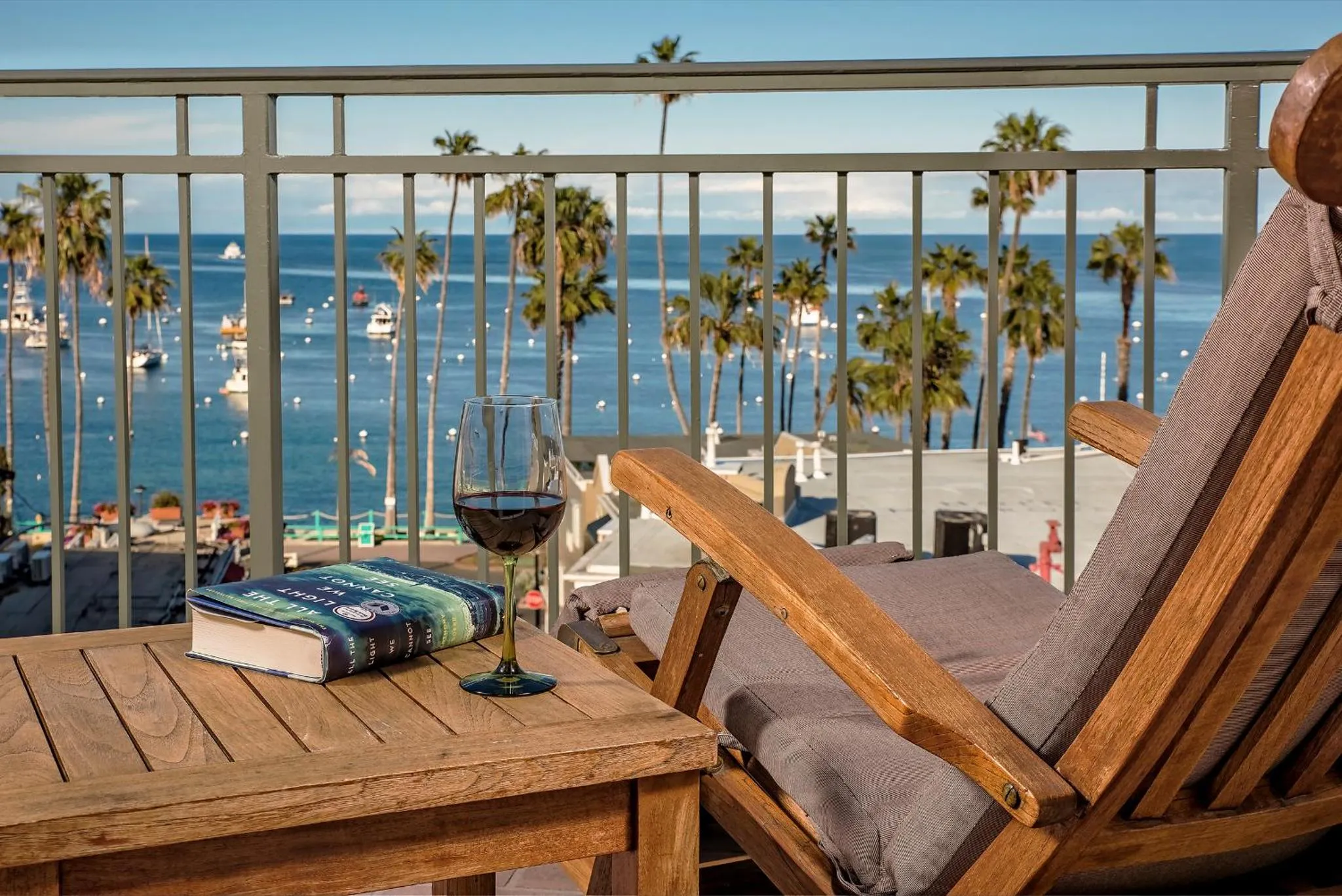 Une terrasse en bois avec une chaise longue, un livre et un verre de vin offre une vue sur la mer et les palmiers.