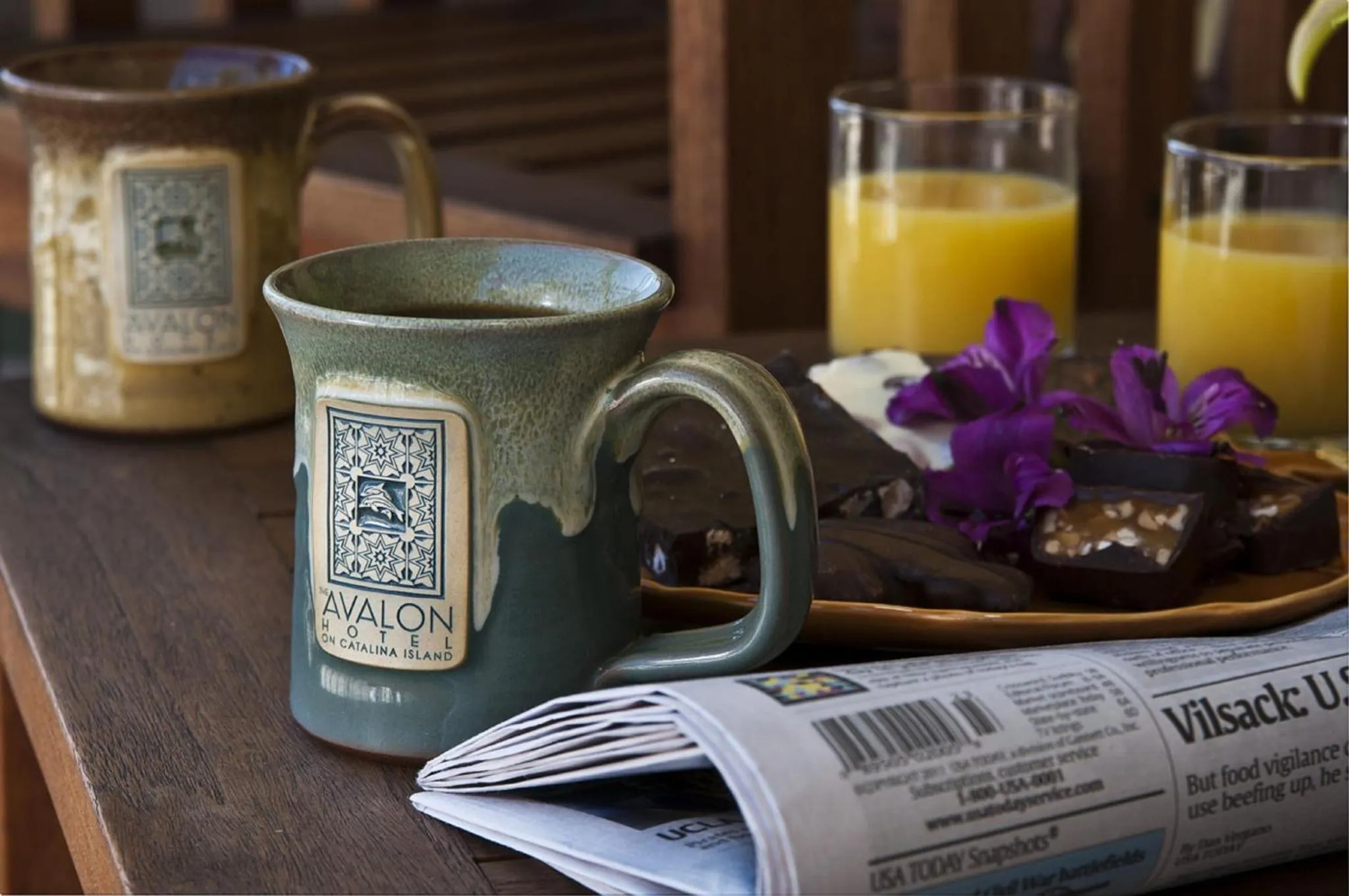 Une table en bois présente des tasses de café, du jus d'orange, une pâtisserie et un journal.
