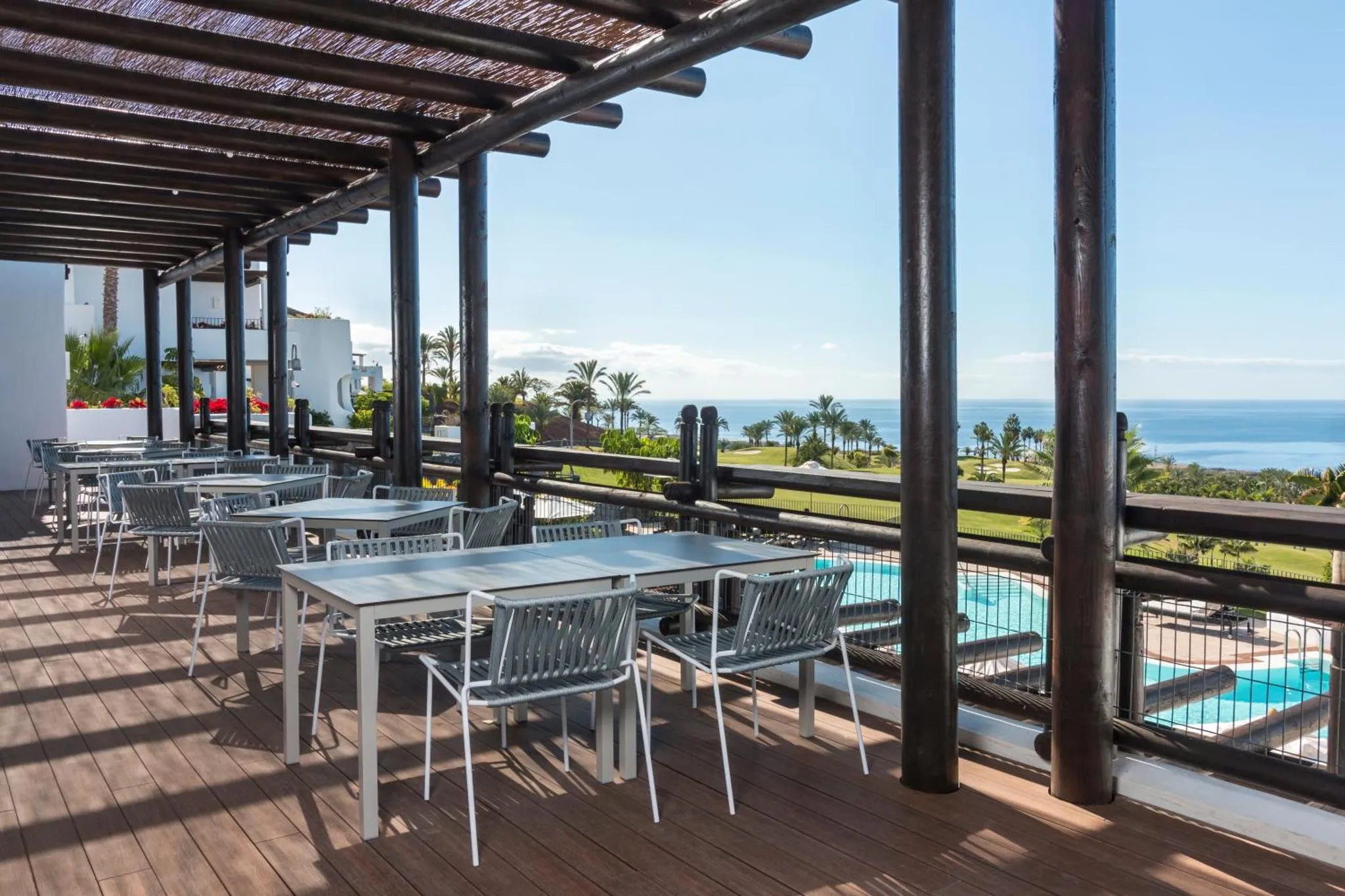 Terrasse ombragée avec tables et chaises donnant sur la mer et une piscine.