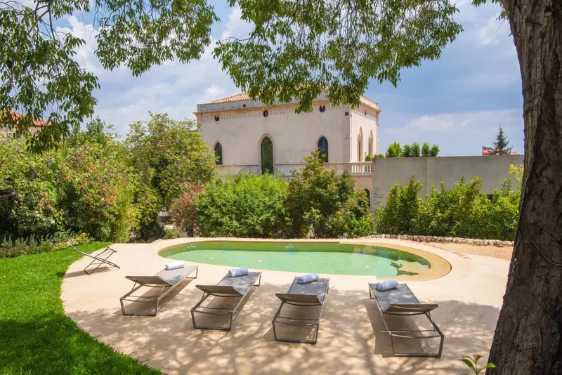 Une piscine rectangulaire bordée de chaises longues se trouve devant un bâtiment blanc à colonnes.