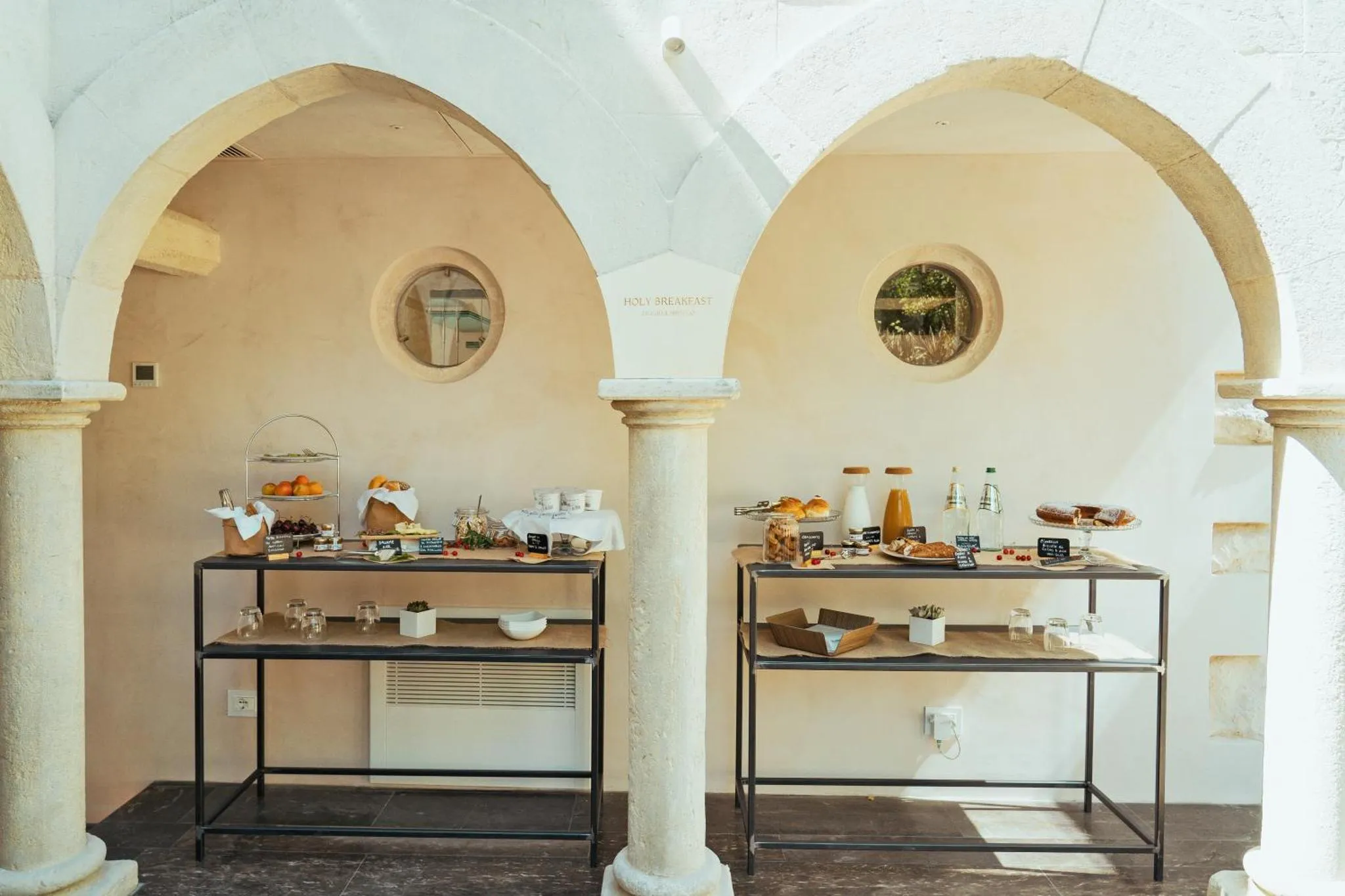Un buffet propose des pâtisseries et des boissons sur des tables en métal dans un espace architectural avec des arches.
