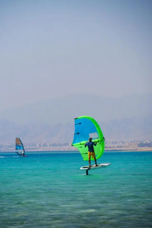 Deux pratiquants de planche à voile naviguent sur une eau turquoise face à des montagnes.