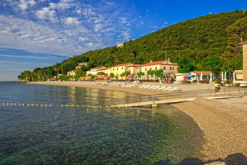 Vue d'une plage de galets bordée de bâtiments et de transats sous un ciel bleu.