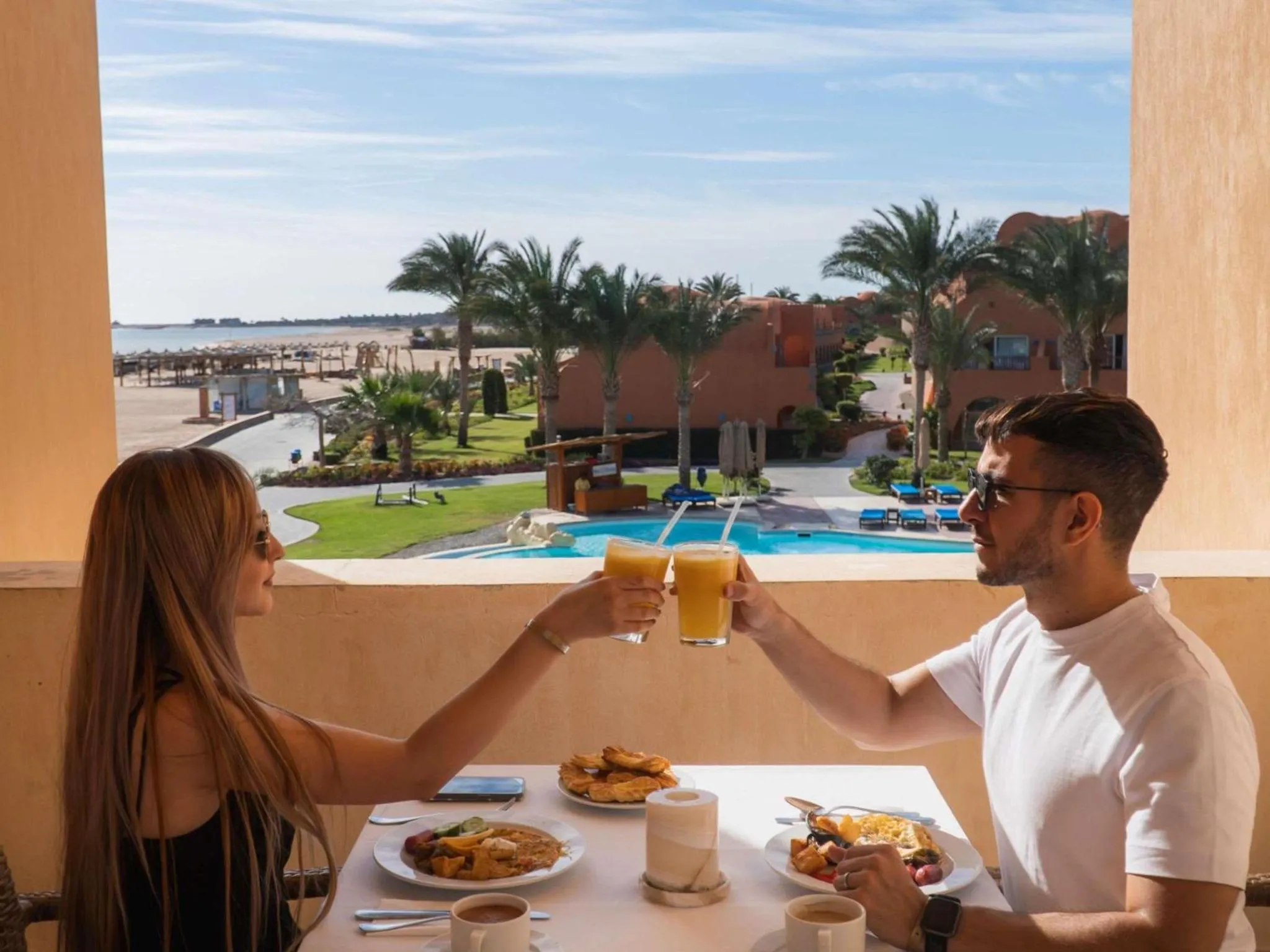 Un couple trinque avec des jus de fruits sur une terrasse ensoleillée avec vue sur la mer et une piscine.