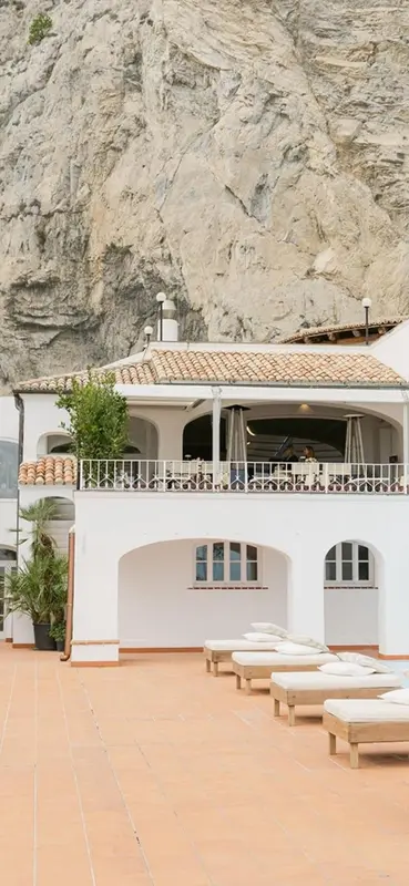 Terrasse avec transats devant un bâtiment blanc et une falaise rocheuse.