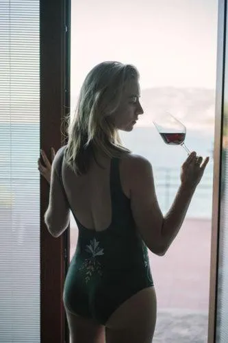 Une femme se tient près d'une fenêtre avec un verre à la main.
