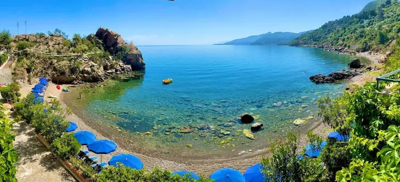 Une plage de galets bordée de parasols bleus s'étend le long d'une crique abritée.