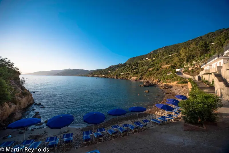Une plage de galets avec des parasols bleus et des transats face à une crique.