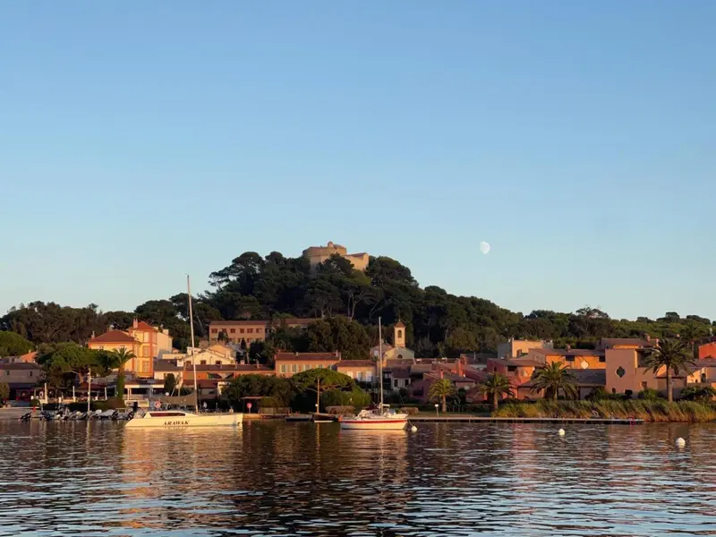 Vue d'un village côtier avec une forteresse sur une colline et des bateaux amarrés dans un port.