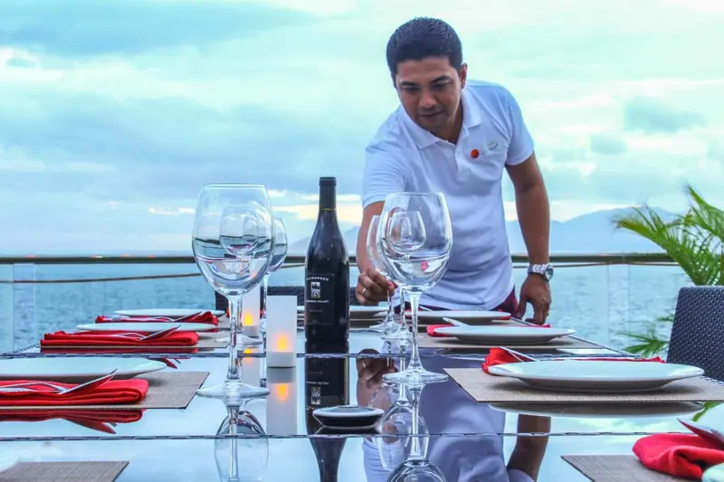 Une table dressée avec des verres et une bouteille de vin, un serveur s'affaire à la préparation.