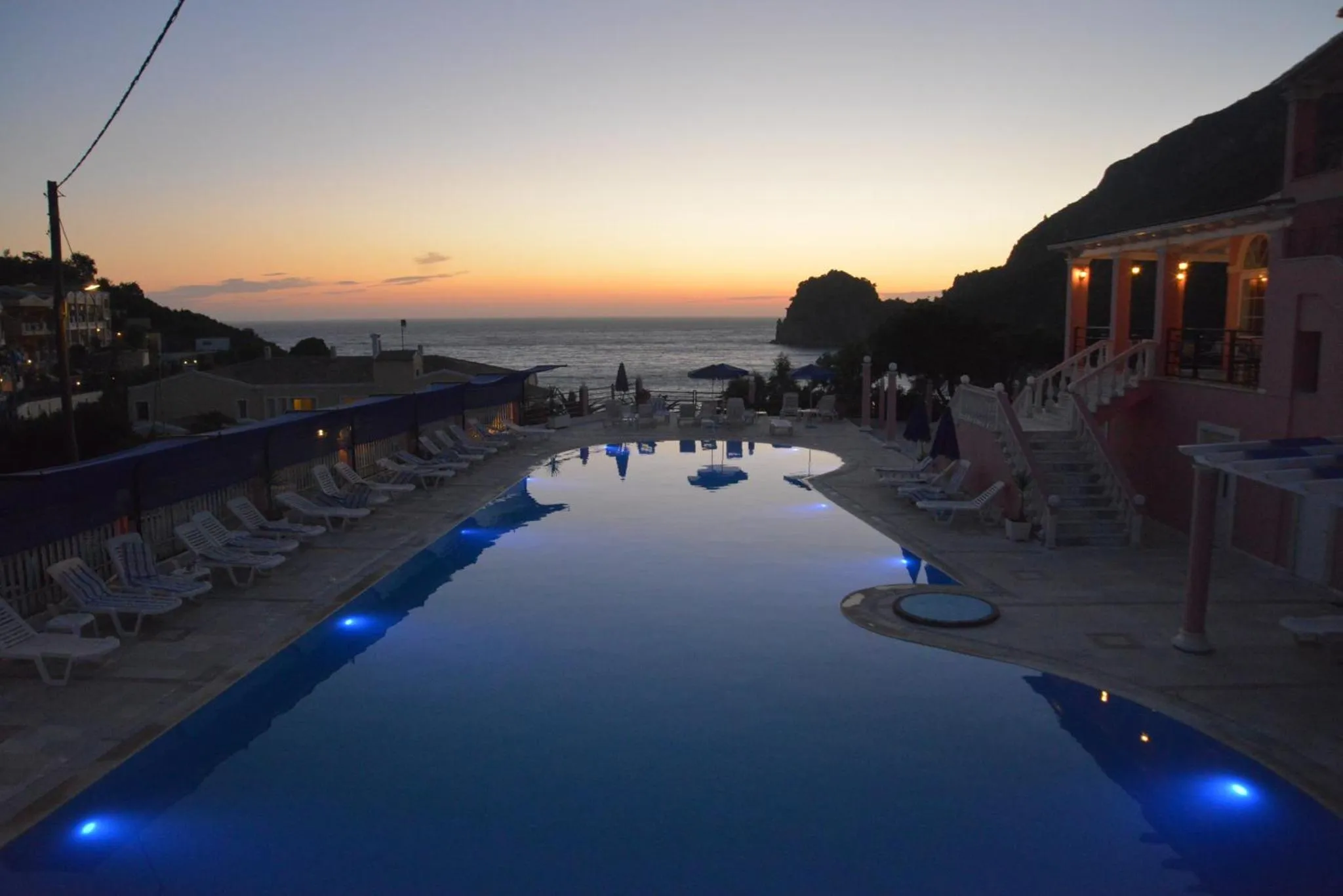 Une piscine illuminée avec des transats au bord d'une terrasse surplombant la mer au coucher du soleil.