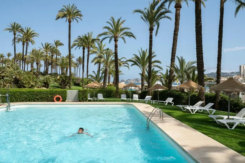 Une piscine rectangulaire bordée de chaises longues et de palmiers sous un ciel bleu.