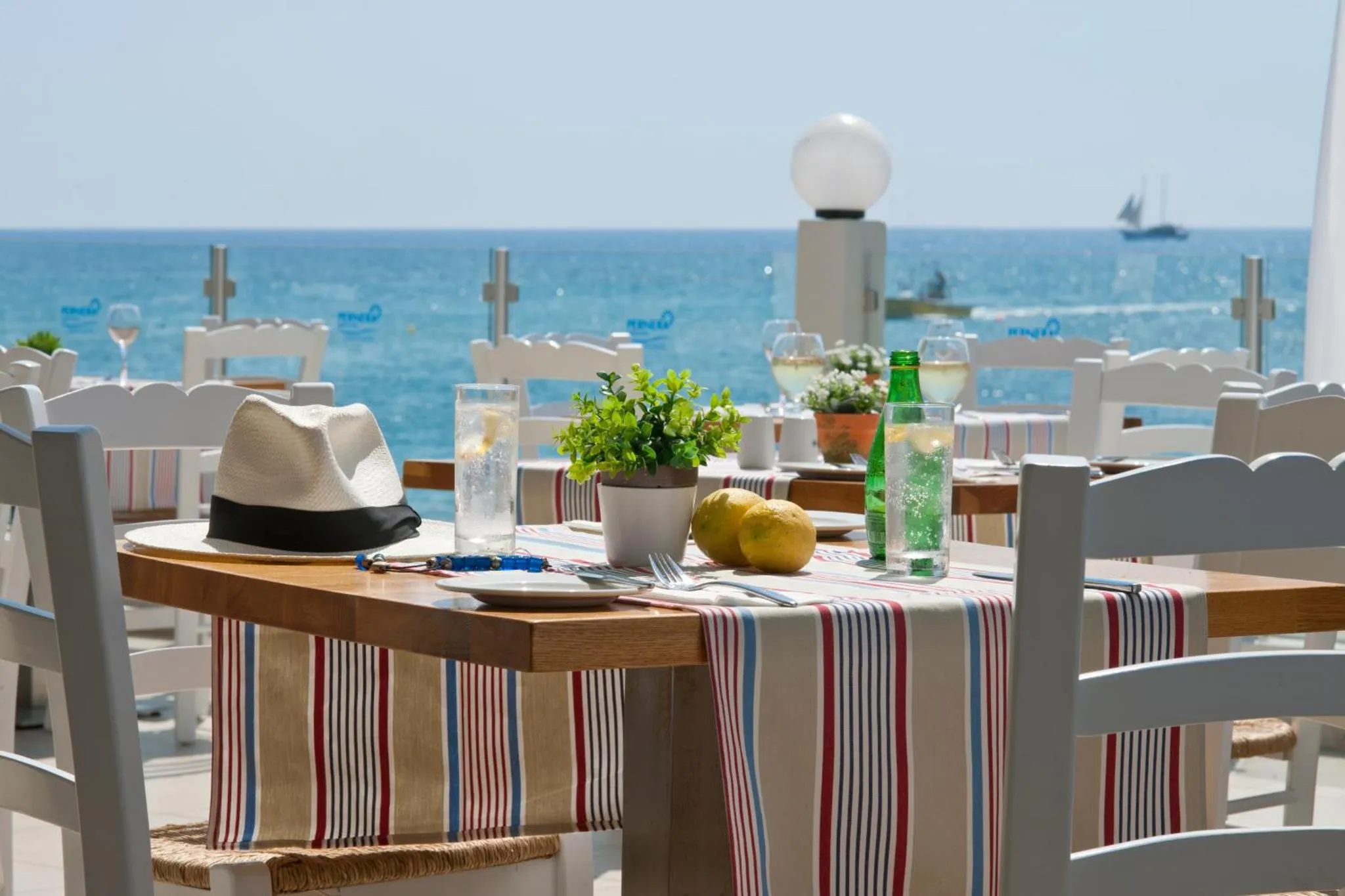 Une table est dressée avec des verres et de la vaisselle sur une terrasse ensoleillée face à la mer.