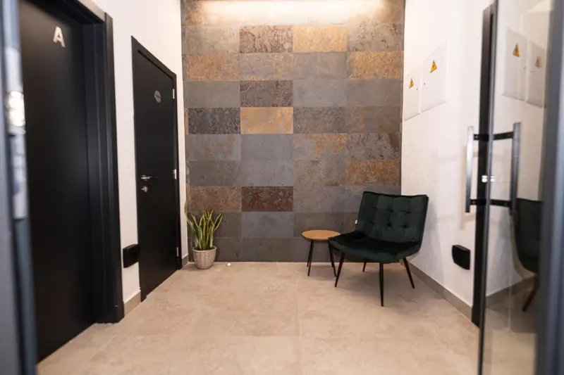 Un couloir d'hôtel mène à des chambres avec un fauteuil et une plante en pot.