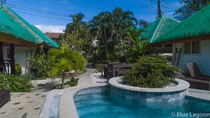 Petites maisons aux toits verts s'ouvrent sur une cour avec piscine et végétation tropicale.