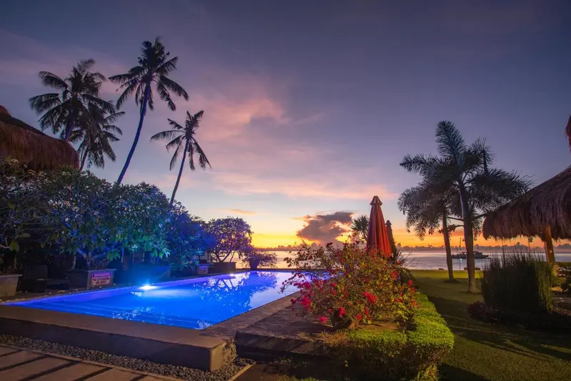 Piscine illuminée au crépuscule avec végétation tropicale et coucher de soleil en arrière-plan.