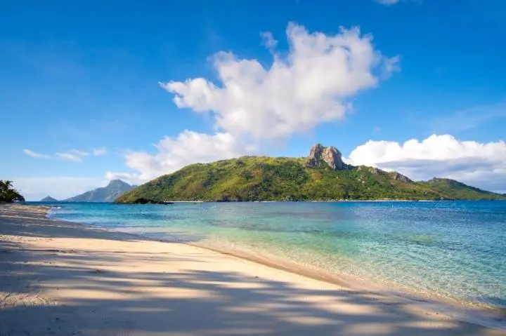 Plage de sable fin bordée d'une eau turquoise et d'une île verdoyante sous un ciel bleu nuageux.