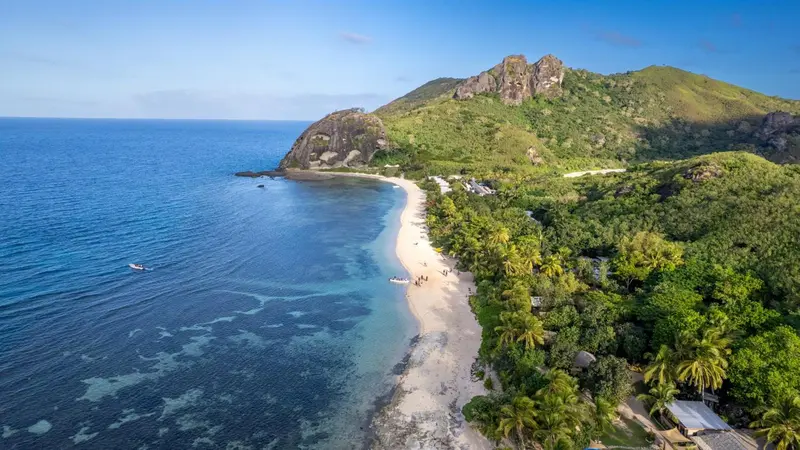 Vue aérienne d'une plage de sable blanc bordée de végétation luxuriante et de formations rocheuses.