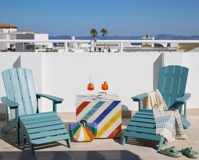 Deux fauteuils en bois bleu turquoise encadrent une table basse colorée sur une terrasse ensoleillée.