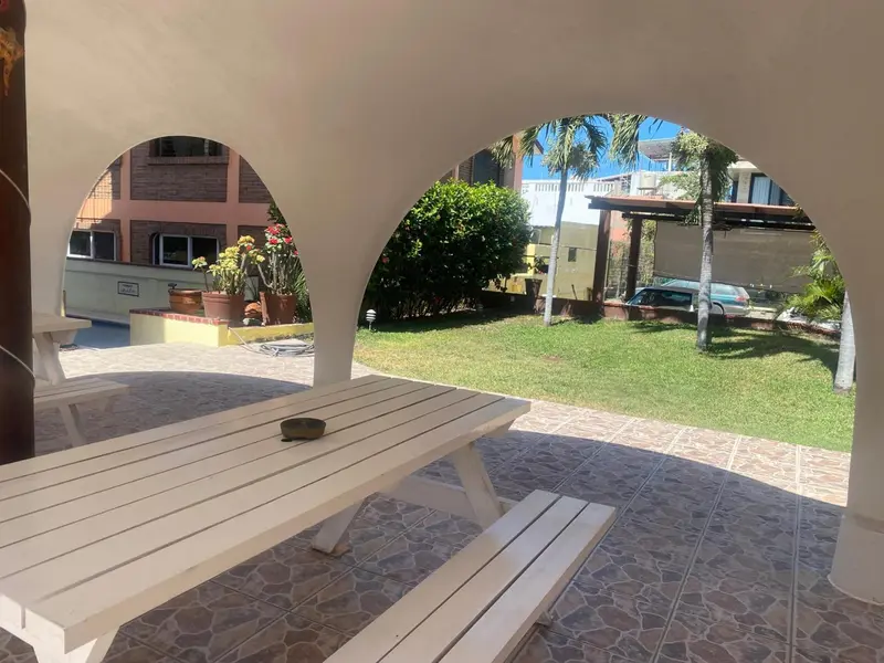 Tables et bancs blancs sous une structure ajourée donnant sur une cour verdoyante.