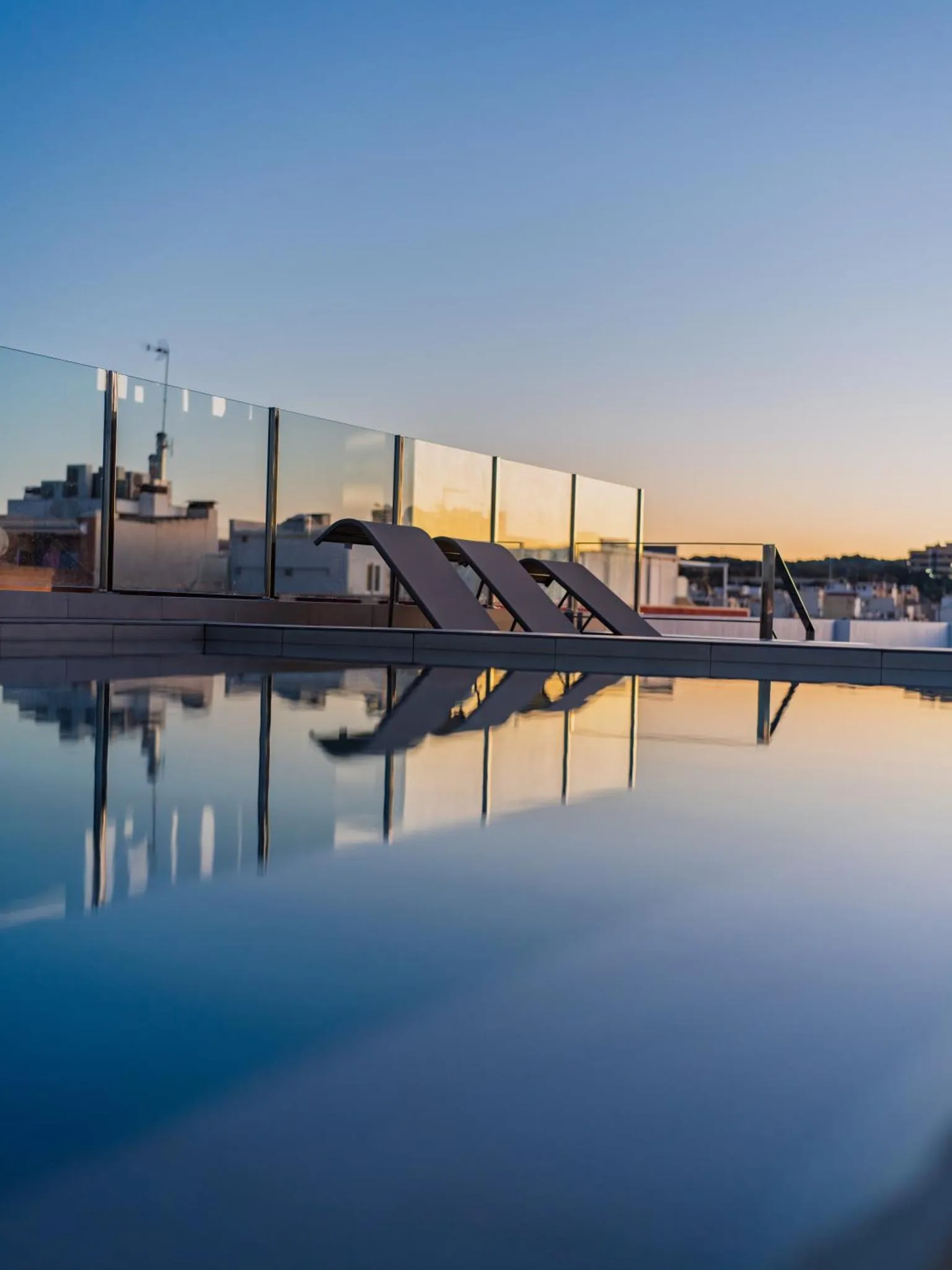 Transats alignés au bord d'une piscine à débordement avec vue sur un paysage urbain au coucher du soleil.