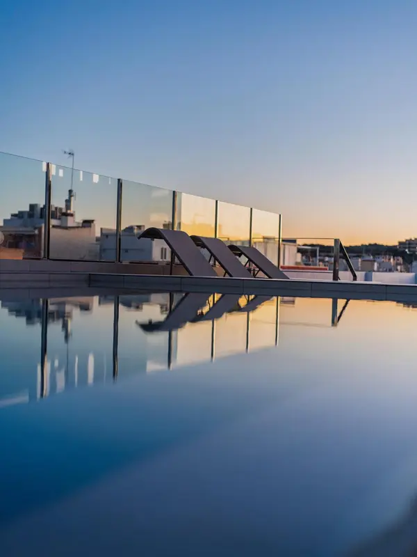 Transats alignés au bord d'une piscine à débordement avec vue sur un paysage urbain au coucher du soleil.