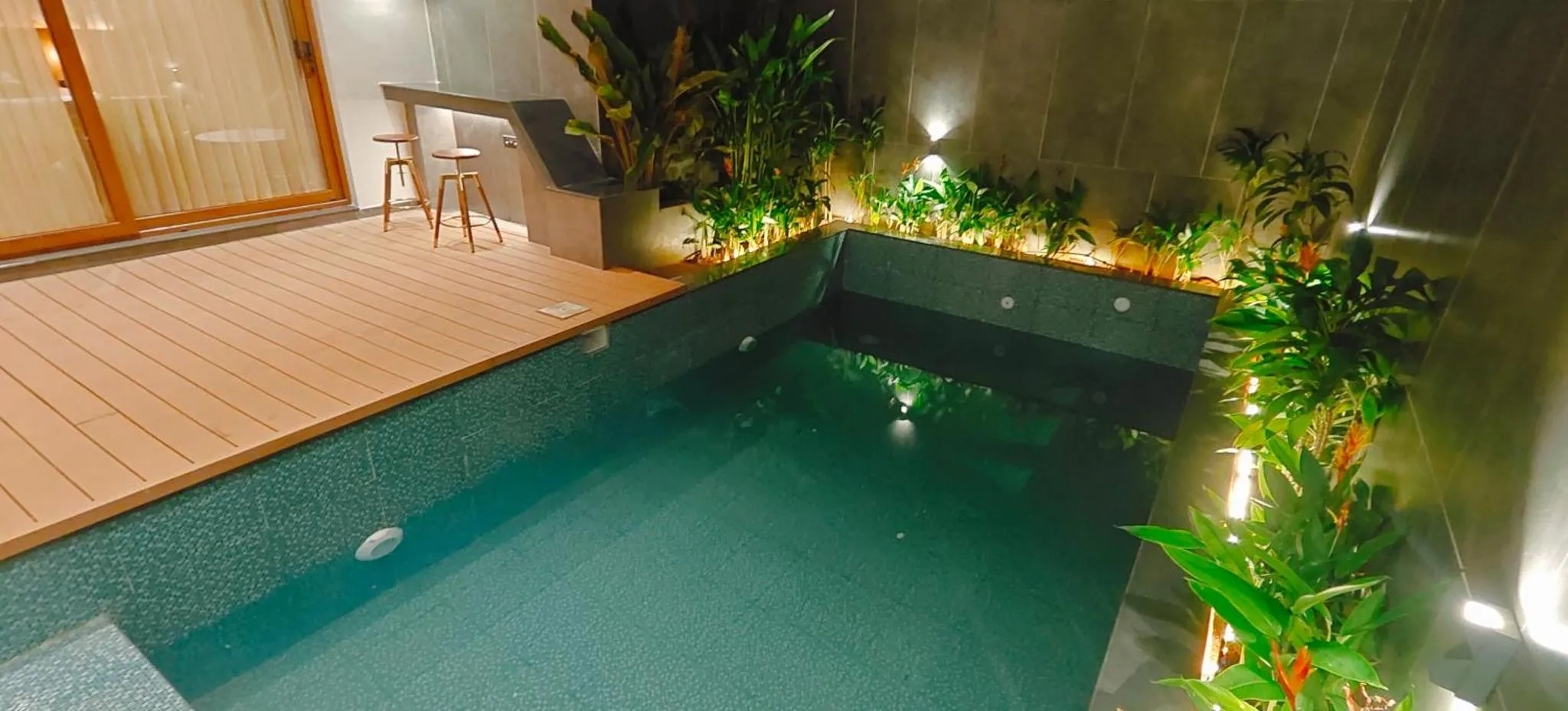Petite piscine rectangulaire avec un espace de détente en bois et des plantes tropicales.