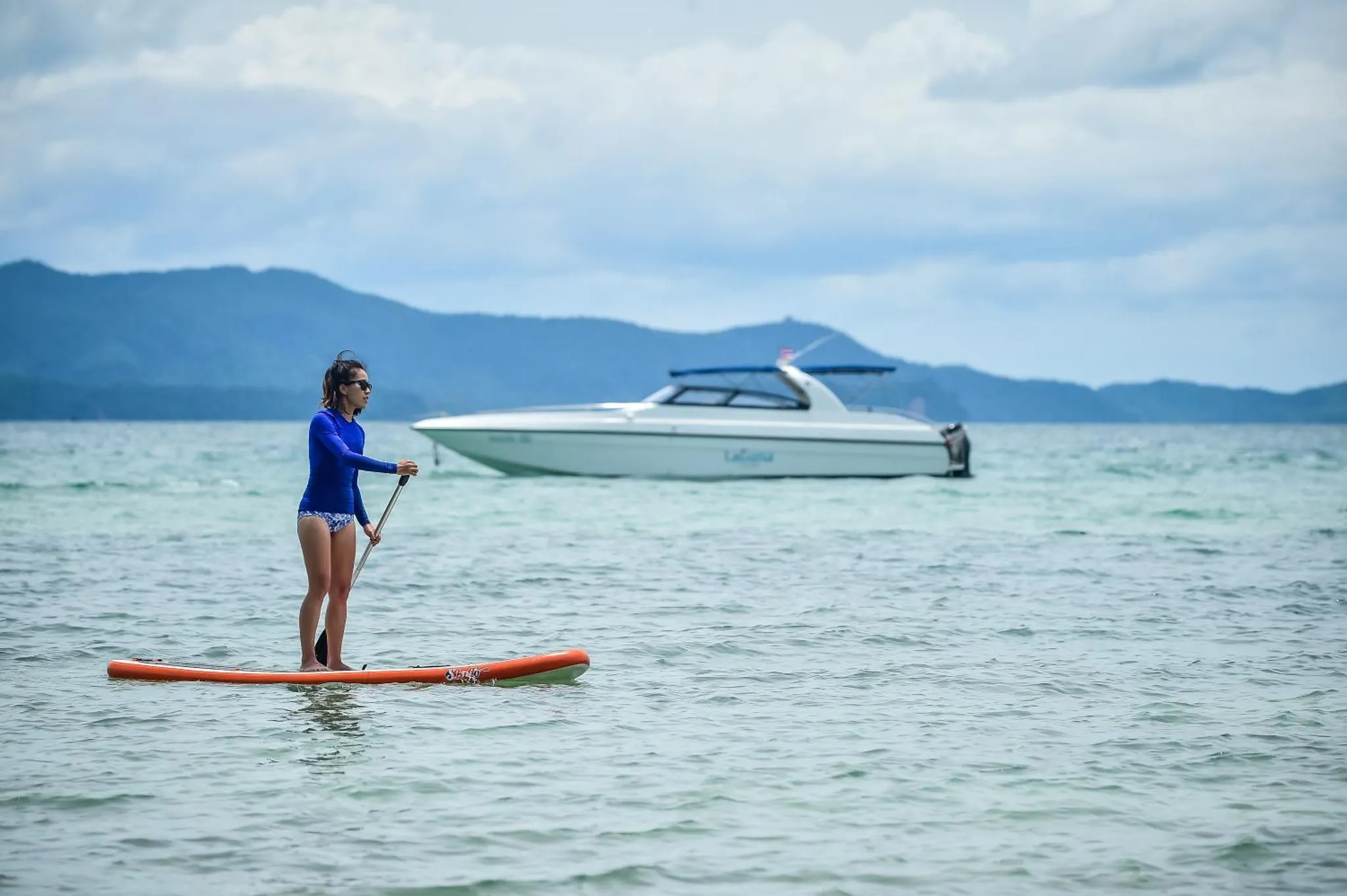 Une personne pratique le stand-up paddle dans une eau turquoise face à une côte montagneuse.