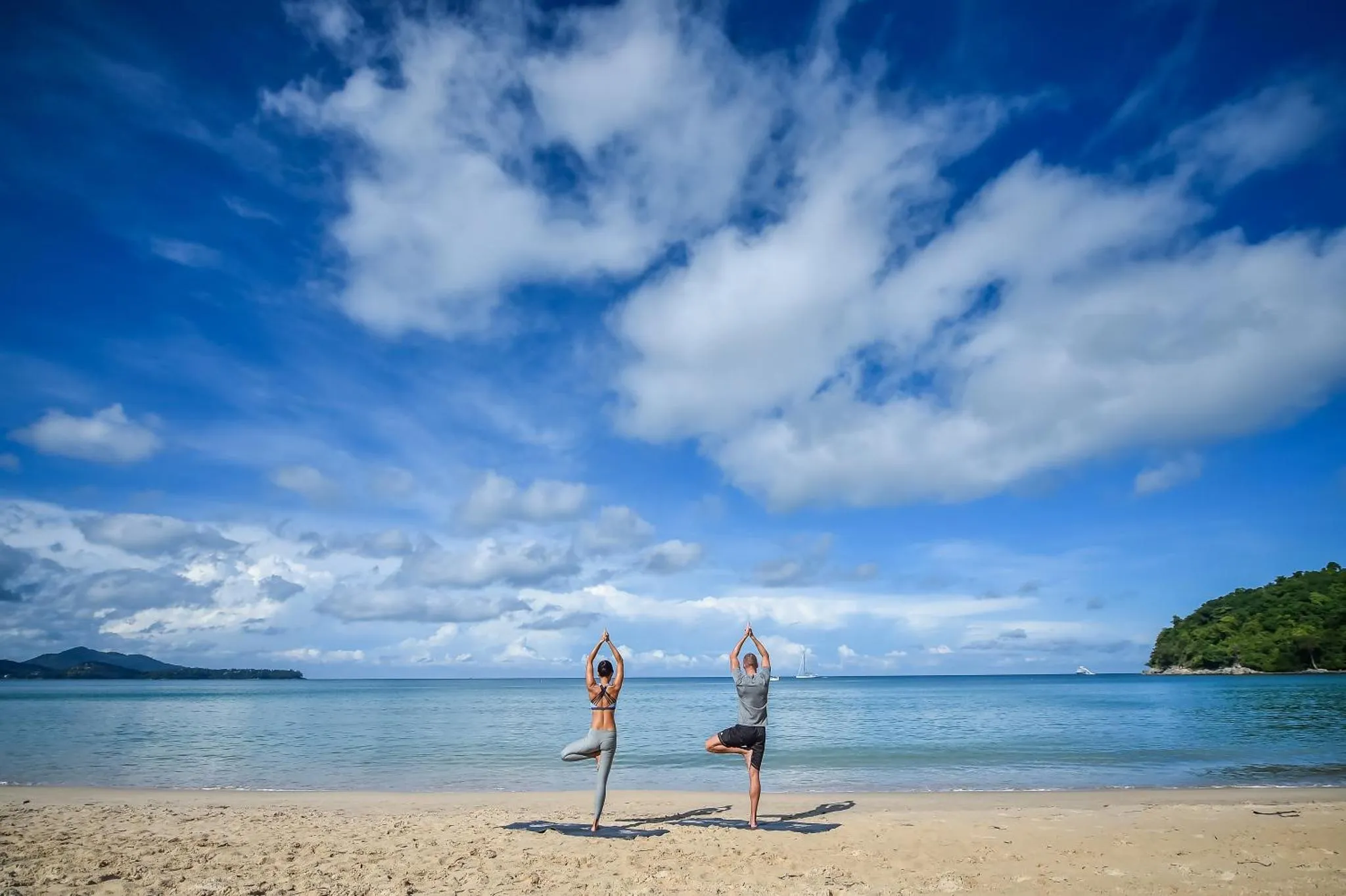 Deux personnes pratiquent le yoga sur une plage de sable fin face à la mer.