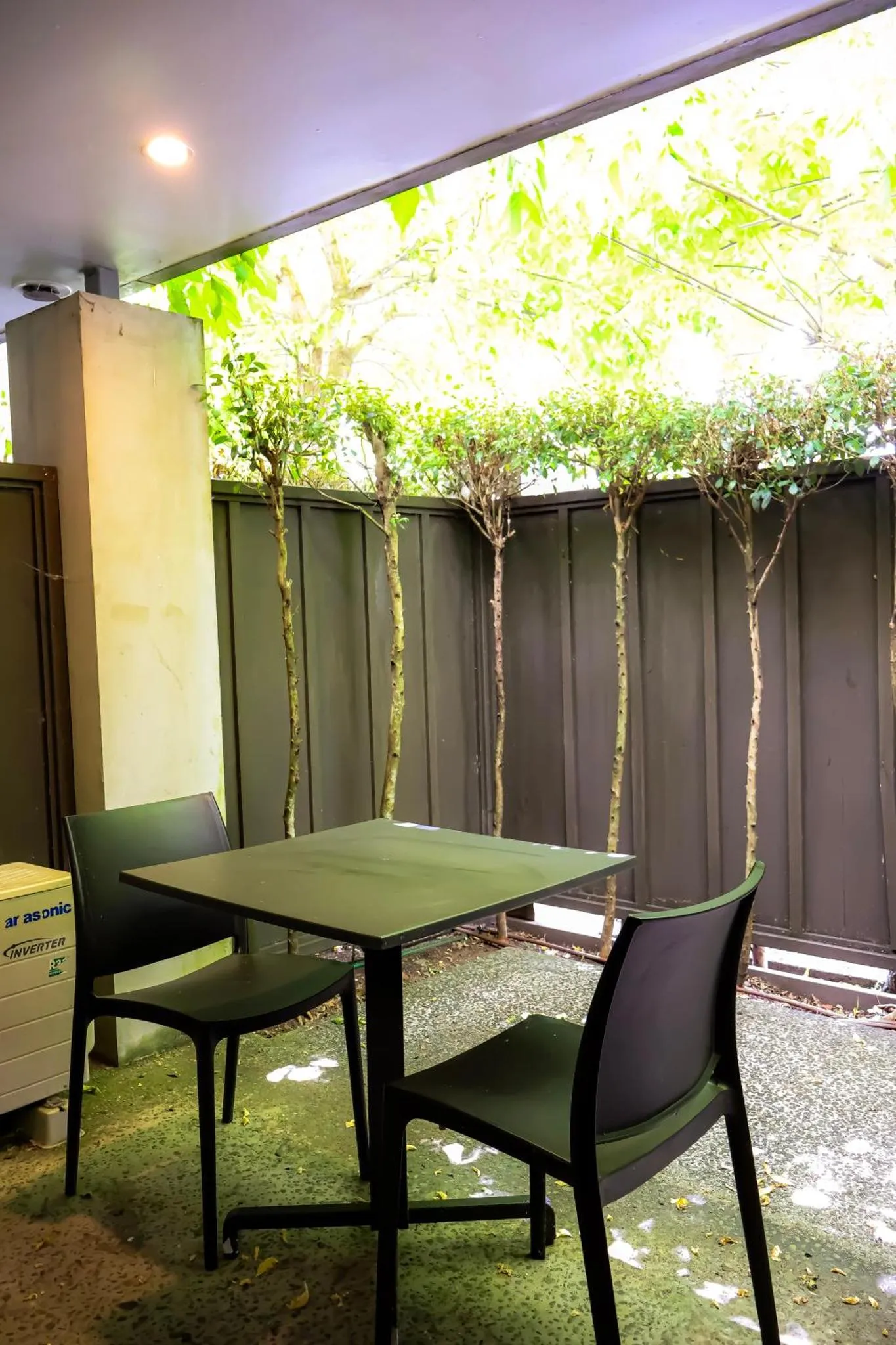 Une table et des chaises en plastique noir sont disposées sur une terrasse ombragée par de la végétation.
