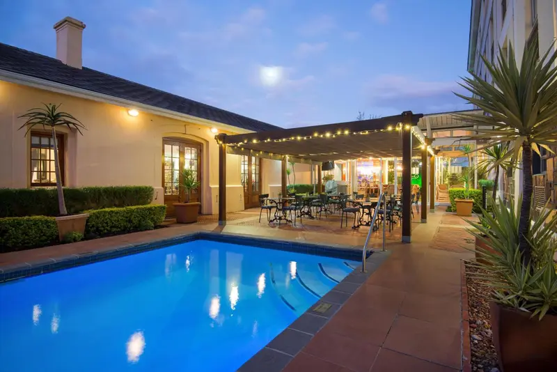 Une piscine rectangulaire au premier plan, avec un espace couvert et aménagé pour les repas à l'arrière-plan.