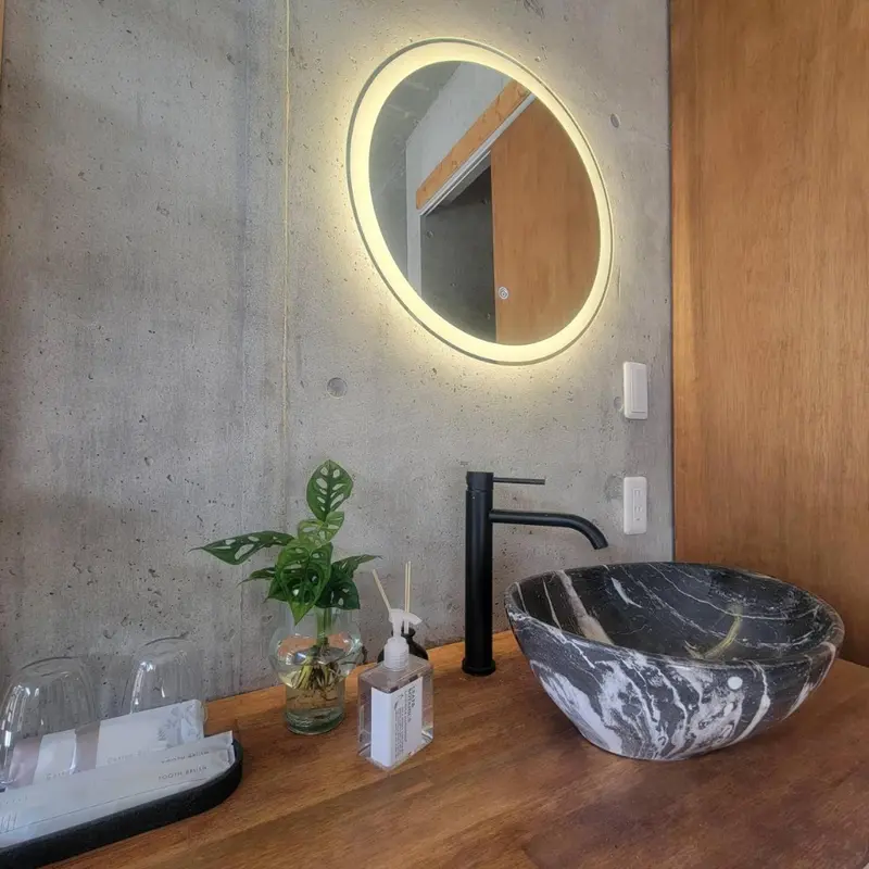 Un lavabo en pierre sombre se trouve devant un miroir rond éclairé dans une salle de bain minimaliste.
