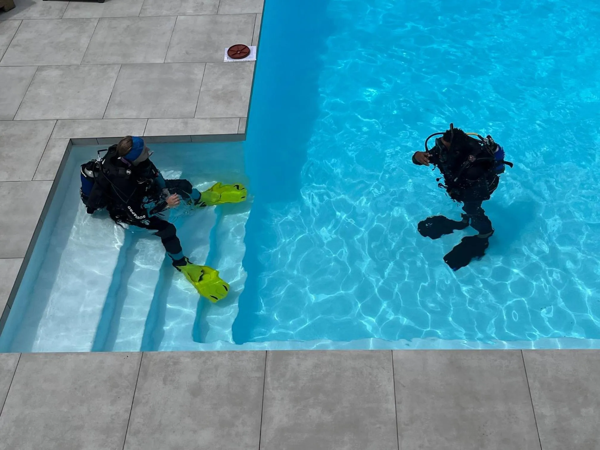Deux personnes en équipement de plongée se tiennent dans les escaliers d'une piscine.