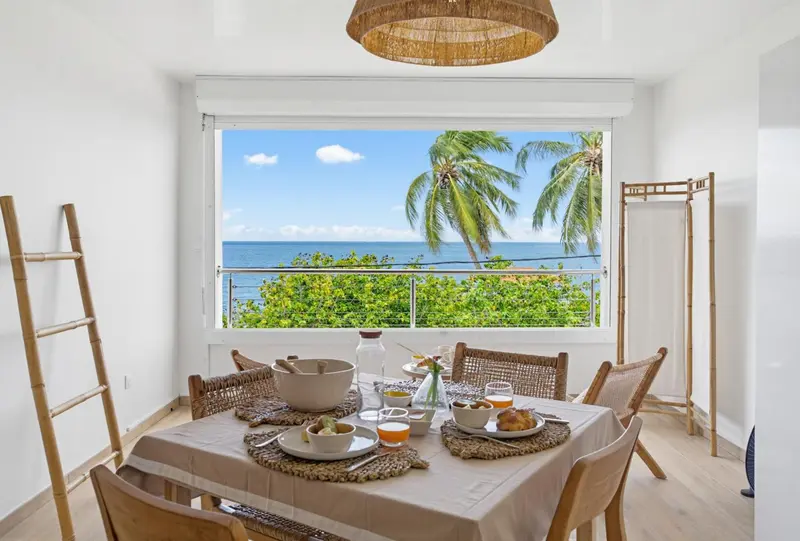 Une table de repas est dressée devant une grande fenêtre donnant sur la mer et la végétation tropicale.