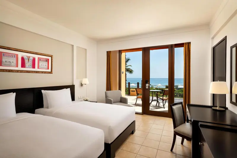 Chambre lumineuse avec deux lits et vue sur la mer par de grandes baies vitrées.