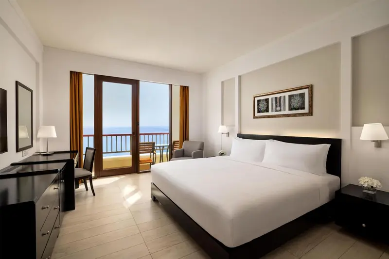 Chambre lumineuse avec un grand lit et un balcon donnant sur la mer.