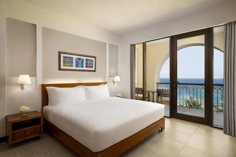 Chambre lumineuse avec un grand lit et une vue sur la mer depuis un balcon.