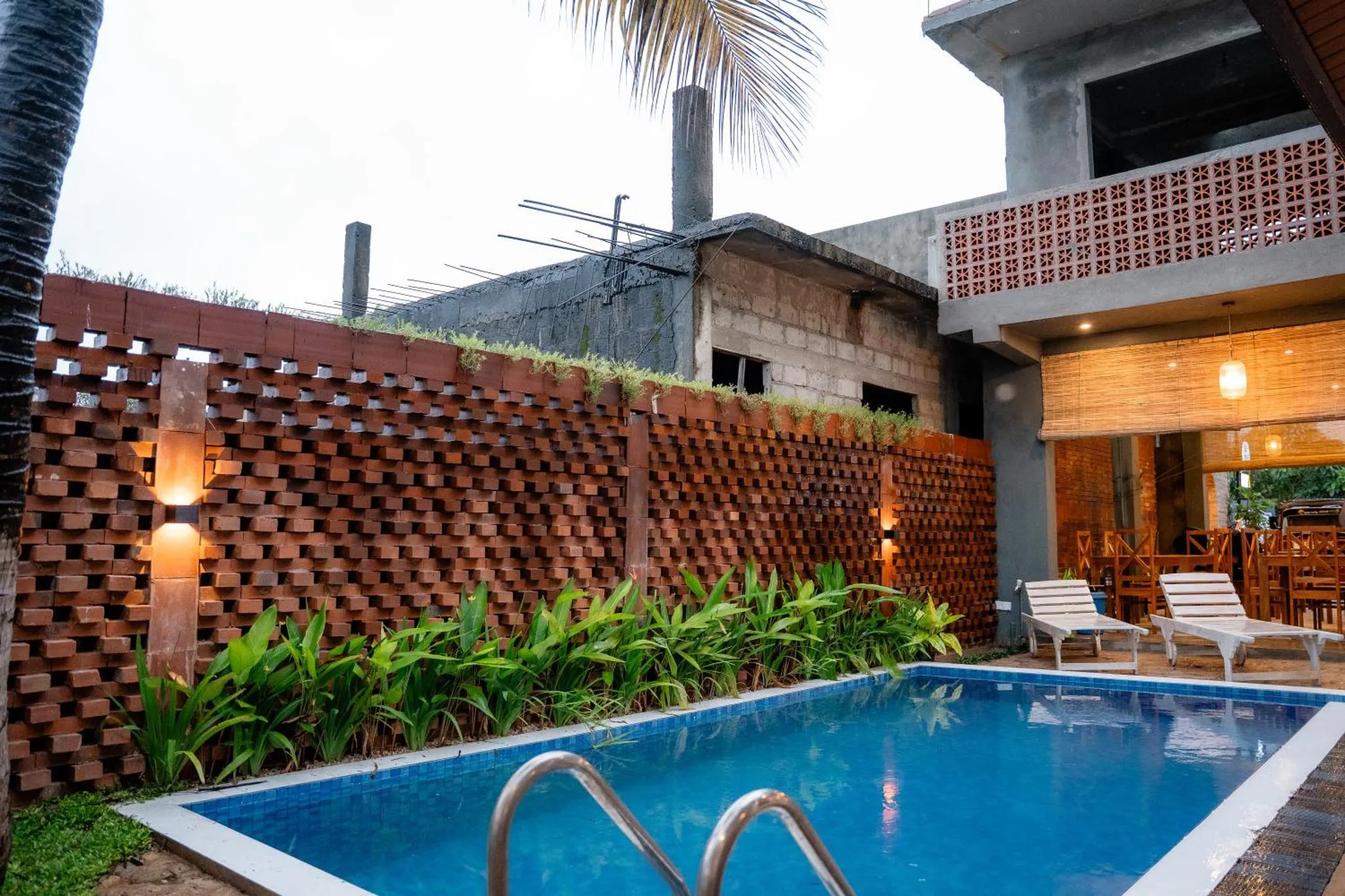Une piscine rectangulaire turquoise est entourée d'une terrasse avec des chaises longues et un mur décoratif en briques.