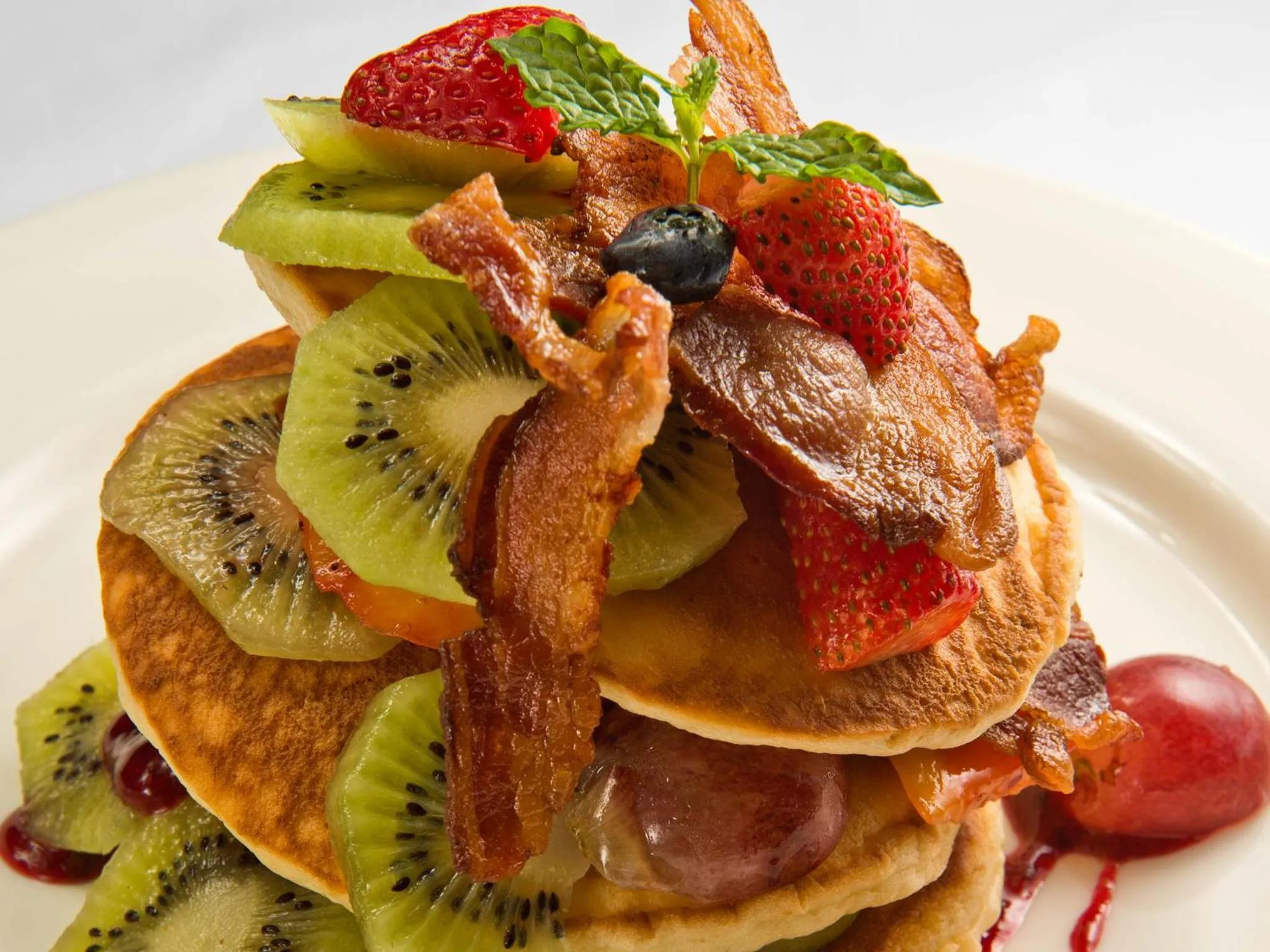 Une pile de pancakes garnis de fruits frais et de bacon croustillant est présentée sur une assiette blanche.